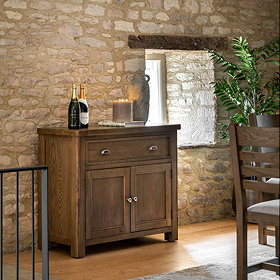 image of dark oak sideboard with one drawer and 2 doors, topped with glasses and pushed against an exposed brick wall