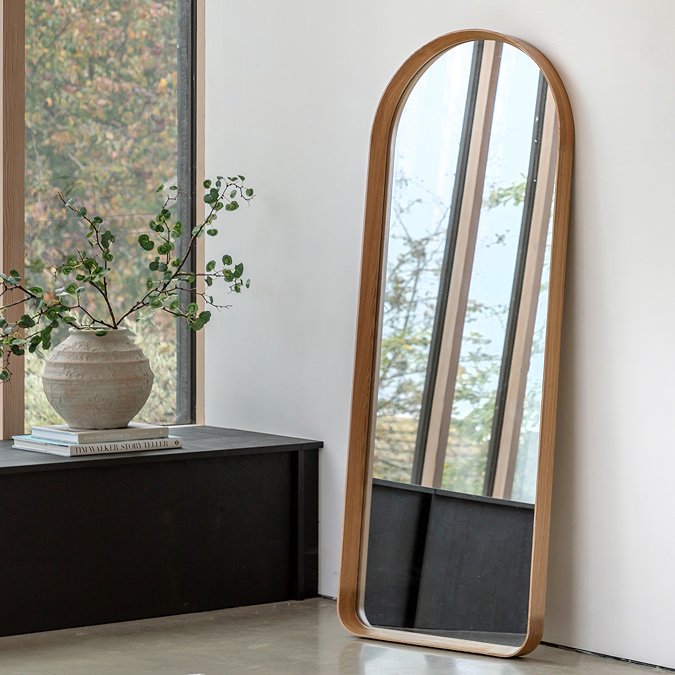 image of floor length rounded rectangular mirror in an oak finish, leaning against a cream wall next to large window