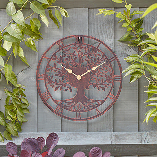 image of bronze wall clock with central tree shape, roman numerals, and gold hands, on a grey garden fence with green foliage around it