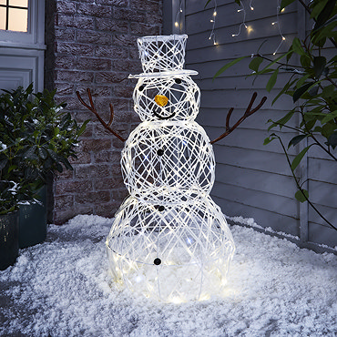 white rattan light up snowman next to white rattan light up reindeer in front of a house, lit up at night on a snowy driveway.