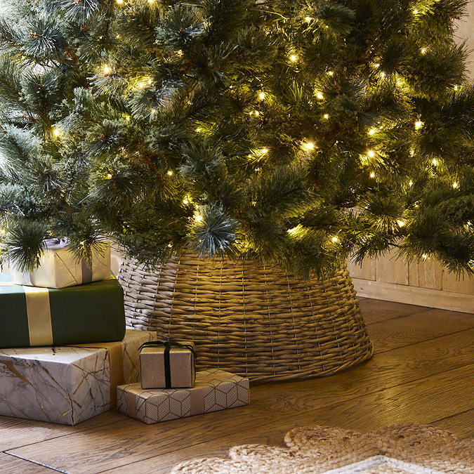 wicker christmas tree skirt at the base of a tree lit with warm white lights, partially obscured by gold-wrapped presents