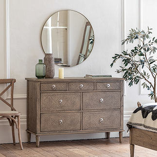 image of seven drawer ashy wood chest, against a wall in a bedroom with round mirror hanging above it