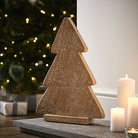angled mage of wooden patterned tree decoration stood on a hearth next to a trio of pillar candles, with christmas tree blurred in the background. 