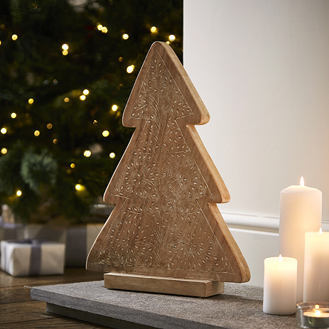 angled mage of wooden patterned tree decoration stood on a hearth next to a trio of pillar candles, with christmas tree blurred in the background.