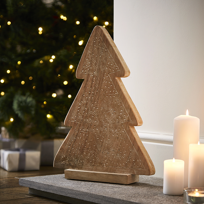 angled mage of wooden patterned tree decoration stood on a hearth next to a trio of pillar candles, with christmas tree blurred in the background. 