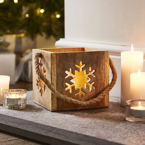 wooden cube tealight candle holder with snowflake design on the side, placed on a hearth next to pillar candles casting a cosy glow