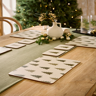set of four placemats with festive woodland trees dotted on them, placed on a warm wood dining table with sage green table runner