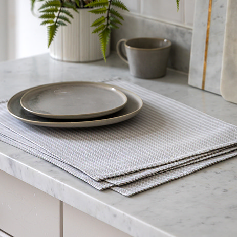 image of four cotton light grey striped placemats on a white marble counter, with two plates on top