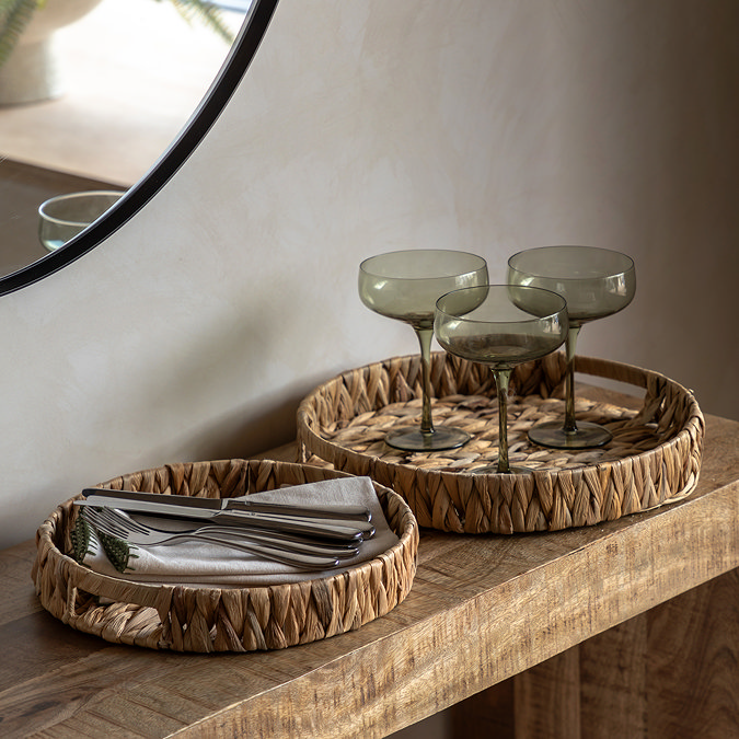 pair of woven round trays, one slightly smaller than the other, displayed on a side table storing cutlery, napkins and glasses.