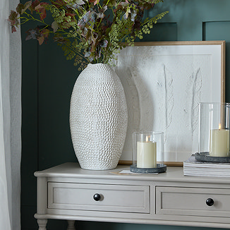 image of tall patterned white ceramic vase on a sideboard filled with faux foliage