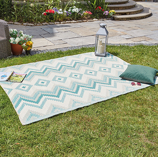 image of green zigzag rug on grass with lantern, cushion, and book on its edge